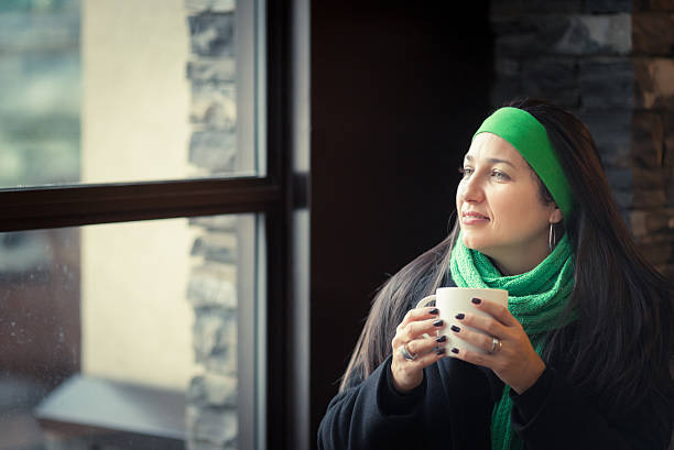 drinking coffee by the window mid adult hispanic woman drinking warm beverage inside and looking thru a window on a winter snowy day. ca04 stock pictures, royalty-free photos & images