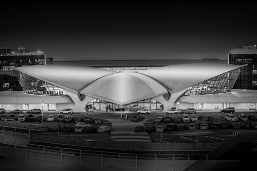 This monochromatic photograph captures the iconic architecture of JFK Airport's Terminal 5 in the evening light. The terminal's sweeping curves and futuristic design stand out against the night sky, a testament to modern engineering and architectural artistry. The dynamic range of lights and shadows casts the structure in a dramatic, almost sculptural relief, inviting viewers to appreciate the interplay between form, function, and design.