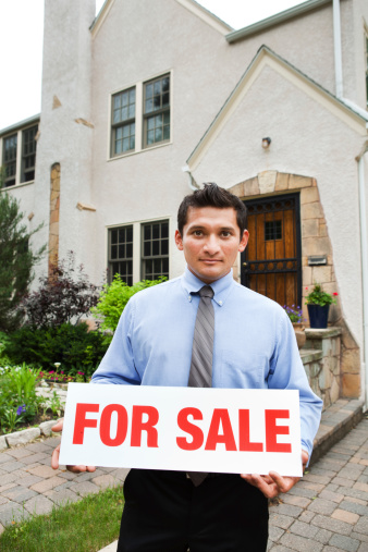 Subject: A serious real estate agent holding up a FOR SALE sign in front of a residential house property.