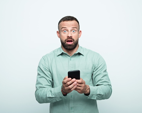 Shocked mid adult men wearing green shirt holding mobile phone in hands and looking at camera. Studio shot, white background.