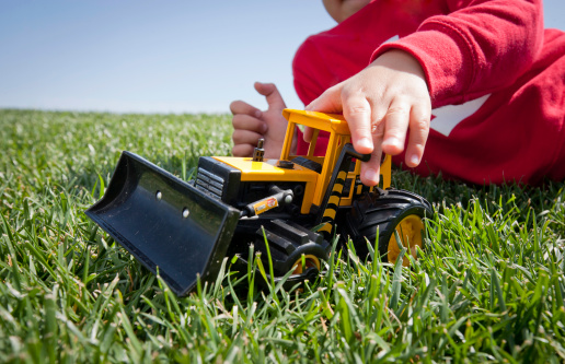 A young boy is playing with a toy construction bulldozer int eh grass while laying down. The cloudless blue sky is visible in the background.