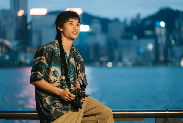 istock Cheerful Chinese young man  holding mirrorless camera sitting at water edge Tsim Sha Tsui evening with Hong Kong Island background 1852394182