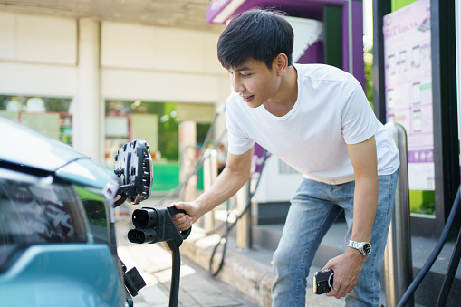 Happy Asian young man using EV charging application on smartphone or mobile phone to access to the charging station. Man plug-in the DC CCS type-2 charging connector to the electric vehicle at public charging station.