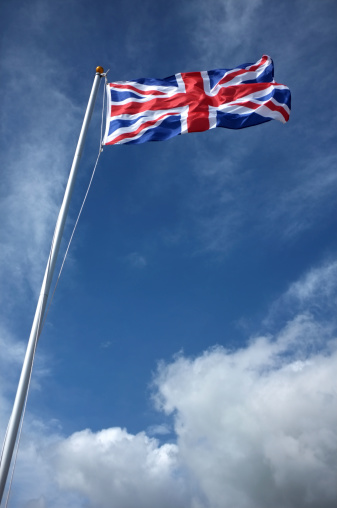 The Union Flag of the United Kingdom flying full mast in full wind against blue sky and stormy clouds.