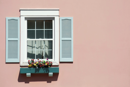 An old sash window with a blue window box and shutters on a pink background. Artificial flowers are in the window box.