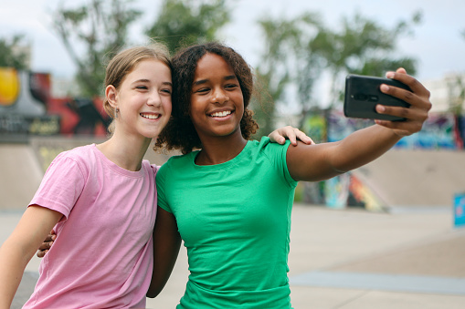 Two friends relaxing after skateboarding, taking a selfie. Both about 12 years old, African and Caucasian females.