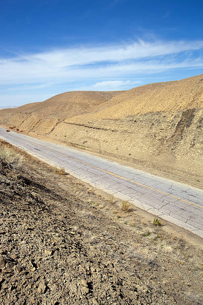 desert badlands road landscape "an old, weathered, cracked two lane country desert road cuts through a desolate and arid desert badlands landscape in the san rafael desert.  vertical composition with copy space in sky taken in green river, utah." sonoran desert desert badlands mesa stock pictures, royalty-free photos & images