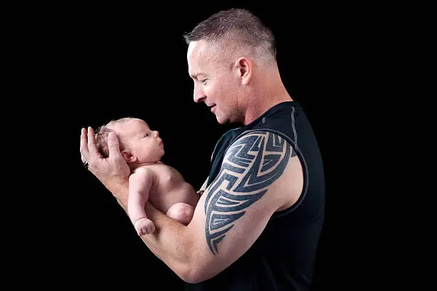 Father with tattoo design on arm holding newborn baby on black background.