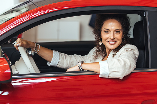 A smiling girl is leaning on the car window and driving while looking at the camera. She got a driving license. A girl driving a car. Travel adventure drive. Portrait of a happy, vehicle and relax female