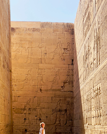 Teenage girl turns her head in shaft of sunlight to admire ancient stone carved hieroglyphics on temple wall in Upper Egypt.