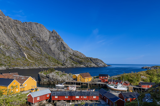 Nusfjord fishing village in Lofoten, Norway, presents a picturesque ensemble of traditional red and yellow wooden Rorbu cabins nestled at the foot of imposing cliffs with the tranquil waters of the fjord reflecting the clear blue sky