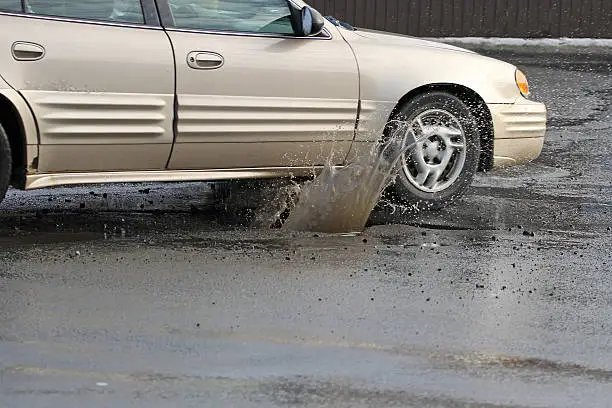 Photo of A champagne colored car hitting a pothole on a rainy day