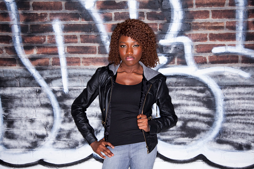 Young black woman (20s) standing in front of brick wall covered with grafitti.