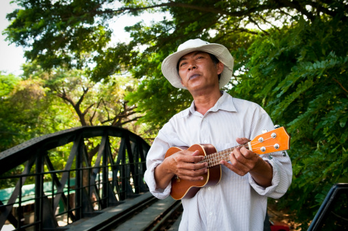 A Thai musician stands and plays his ukulele on the famous Bridge on the River Kwai in Kanchanaburi, Thailand.