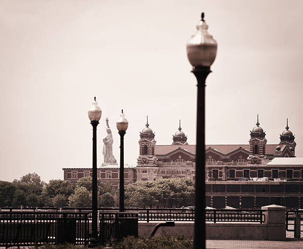 Desaturated view of the Statue of Liberty between and beyond several old-fashioned street lamps. Photo taken from Liberty State Park in Jersey City, New Jersey, USA - looking south across Ellis Island. The Statue of Liberty stands on Liberty Island near the mouth of the Hudson River in the Upper Bay of New York harbor. The Ellis Island Immigration Museum at center right is being refurbished in this photo from May 2011.