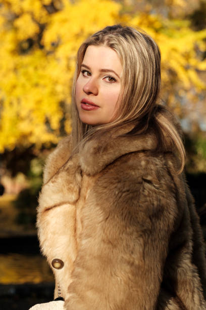 Young caucasian blonde woman is posing in a park in front of autumn yellow background. She is looking at camera and she is wearing a winter faux fur coat.