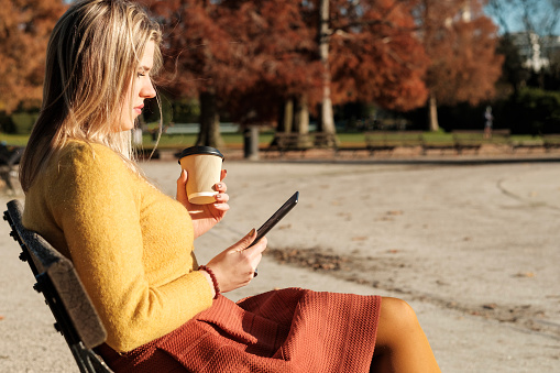 Young woman sitting on a bench park and reading on an E-reader while she is holding a cup of coffee. It is autumn or winter and she is wearing a yellow sweater and orange wool skirt.