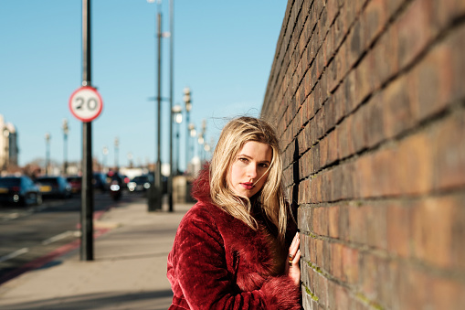 Portrait of young woman wearing a red fake fur coat leaning against a brick wall. She is beside a traffic busy road and it is a sunny autumn or winter day.