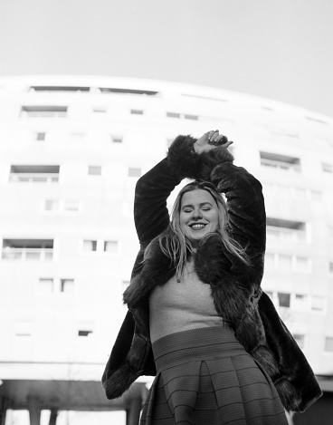 Smiling female model posing with white building behind in a cold winter day. Her arms are up and the image is monochromatic, made with an old film camera.