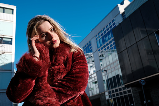 Serious portrait of proud young woman posing between modern buildings. She is looking straight to camera and she is wearing a red fake fur coat in a very cold winter day.