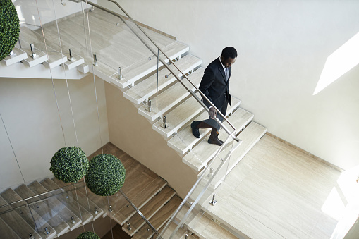 High angle view at Black man in business suit going downstairs holding on handrail in modern office building, copy space