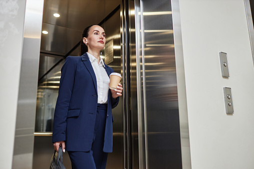 Low angle view at Caucasian brunette businesswoman holding coffee cup and bag dressed in suit leaving elevator in office building, copy space