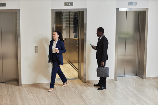 Wide shot of African American businessman holding phone and bag looking at Caucasian business lady exiting elevator in office building
