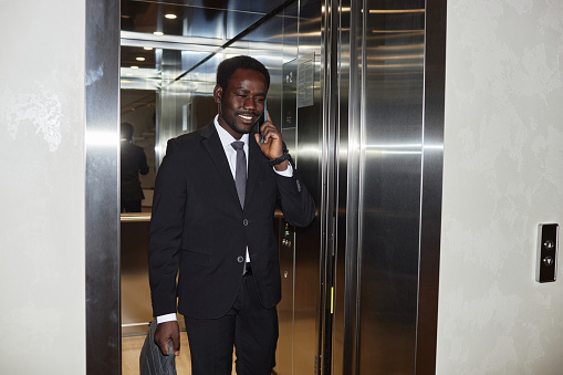 Medium full shot of smiling Black man wearing elegant business suit talking on smartphone while leaving office elevator, copy space