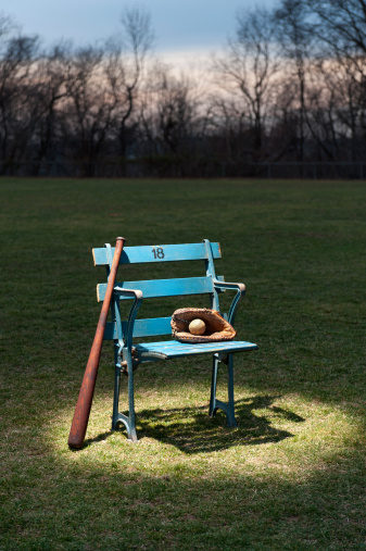 An antique baseball stadium seat from the 1920's spot lit outside on a field at night. There is a glove and bat with the chair.
