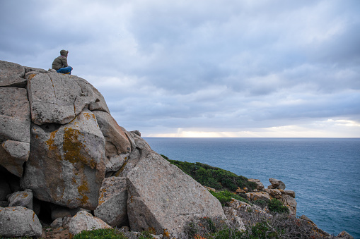 Young man sits on rock bluff above sea on stormy day