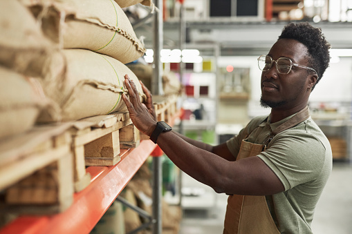 Side view portrait of Black young man inspecting burlap coffee bags on shelf in roastery