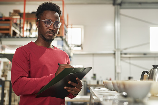 Waist up portrait of Black young man as coffee expert taking notes on clipboard during cupping and quality control at coffee factory, copy space