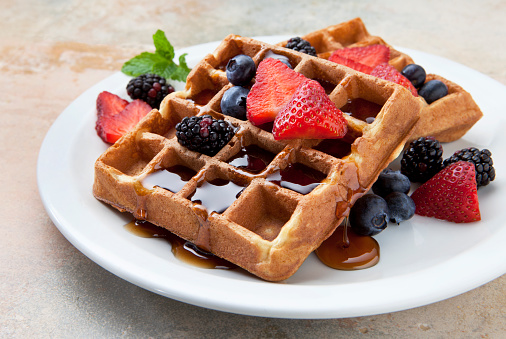 Two square Belgian waffles on a white plate on a marble counter. The golden brown waffles are topped with blueberries, strawberries and blackberries. Maple syrup pools in the waffle pockets and overflows onto the plate which is garnished with a sprig of fresh mint.