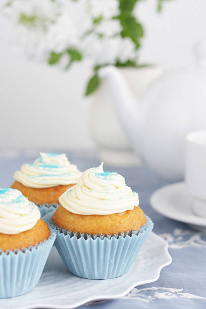 A studio shot of cupcakes served for an afternoon tea. A white plate with wave edges stands in the foreground on a table dressed with a vintage blue tablecloth. The plate holds three cupcakes with vanilla buttercream in pale blue cupcake liners. A white tea pot and tea cup stands at the background. A vase with white flowers stands behind. Shallow depth of field.