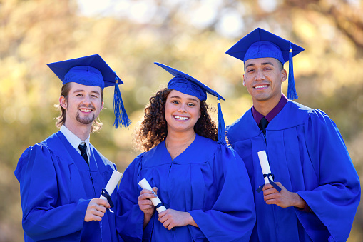Three classmates wearing bright blue cap and gown holding diplomas.