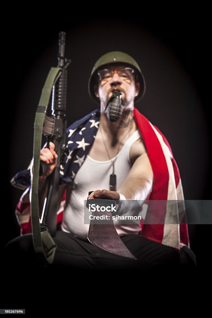 Soldier Wearing American Flag, Holding Grenade in Mouth, Machete "A soldier wearing black pants, a white tank top, a green combat helmet, glasses, and an American flag across his shoulders poses while sitting against a black background. He is holding a grenade with his teeth. In his right hand is a rifle, in his left is a machete, which is pointing at the camera, and holds focus. He faces the camera with a casual, confident pose." 30-39 Years Stock Photo