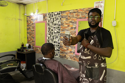 A barber in his barbershop holds a razor while his customer looks at himself in the mirror.
