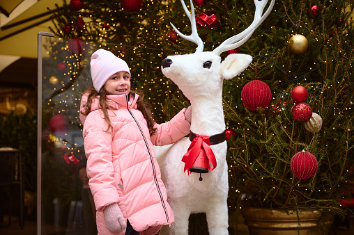 Cheerful lovely Caucasian smiling happy child girl enjoying the upcoming winter holidays, standing against a Christmas tree near a white deer. December 25. Happy New Year and Merry Christmas