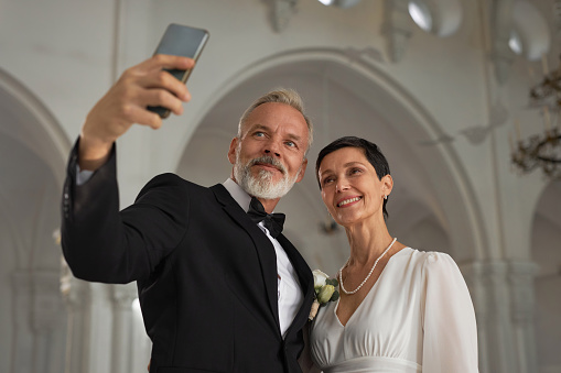 Waist up portrait of senior bride and groom taking selfie photo together via smartphone in church interior, copy space