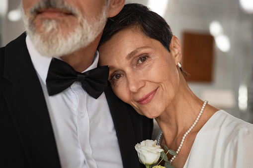 Close up portrait of beautiful senior woman as bride laying head on grooms shoulder and smiling at camera
