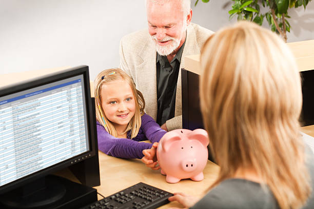 Child Handing Coin Piggy Bank, Opening Bank Account with Teller A cheerful grandparent helping a smiling young girl grandchild with her piggy bank savings. Opening a children bank account with the bank teller in a retail bank counter. Photographed indoors in horizontal format. bank teller stock pictures, royalty-free photos & images