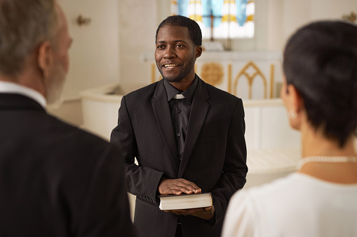 Waist up portrait of smiling Black young man as priest officiating wedding in church and holding Bible