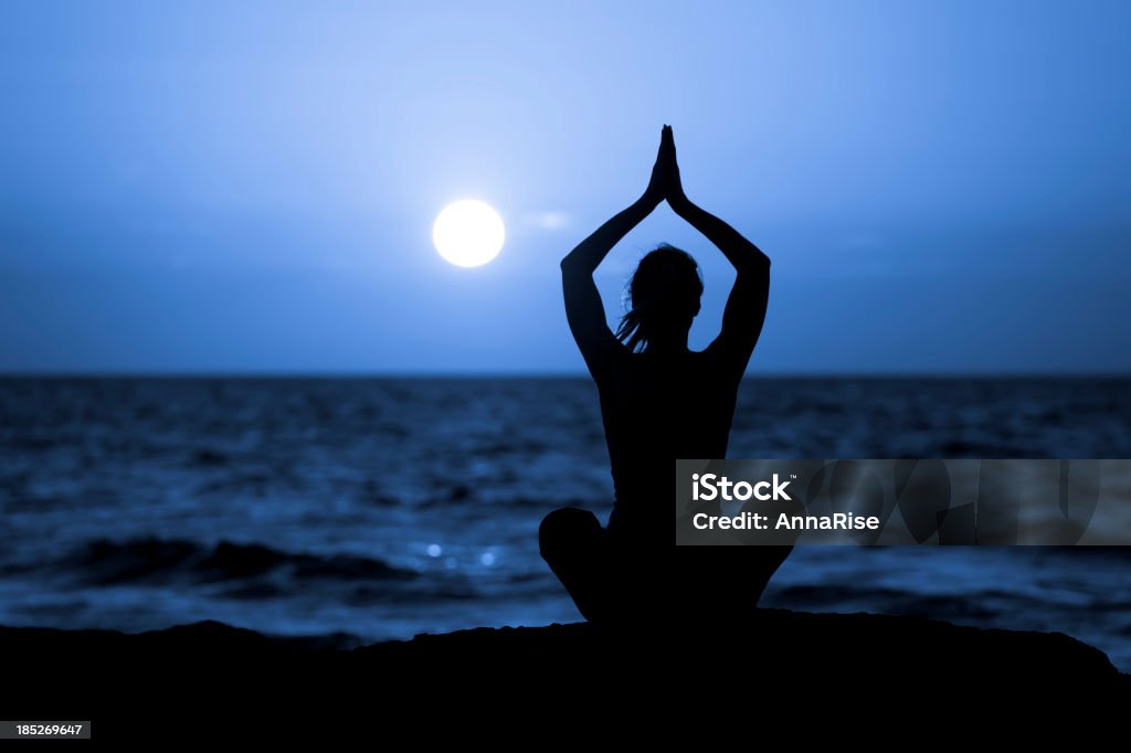 Silhouette of Young Woman Doing Yoga Silhouette of woman in lotus position sitting on the beach and medditating with her hands raised. Moon Stock Photo