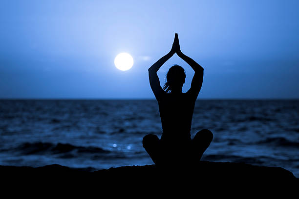 Silhouette of Young Woman Doing Yoga Silhouette of woman in lotus position sitting on the beach and medditating with her hands raised. mantra stock pictures, royalty-free photos & images
