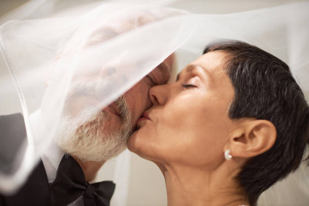 Close up portrait of loving senior bride kissing husband on cheek on wedding day covered with veil