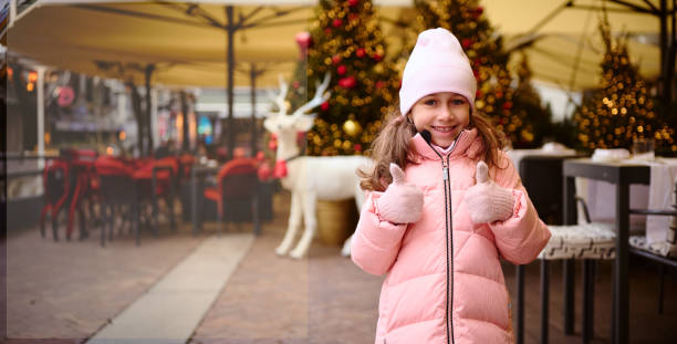 Happy kid girl thumbs up and smiles cutely looking at camera, expressing joy and positive emotion. New Year concept Happy kid girl in warm clothes and woolen mittens, standing in the city at Christmas fair, thumbs up and smiles cutely looking at camera, expressing joy and positive emotion. New Year and Xmas concept cutely stock pictures, royalty-free photos & images