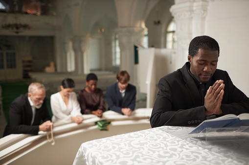 Portrait of young African American priest praying at altar with group of people during Sunday service in church