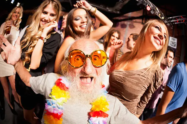 An older man wearing large yellow-tinted eyeglasses is dancing and laughing, with younger people dancing behind him.  The image is in a fish eye view with the focus on the older man.  There are bright lights hanging on a black ceiling of the type found in dance clubs.