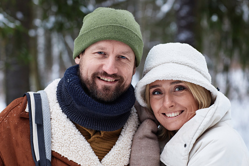 Affectionate smiling couple in winterwear standing in front of camera and looking at you in the forest with evergreen trees covered with snow