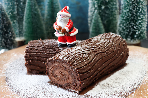 Stock photo showing close-up view of a chocolate Yule log cake that is prepared as part of the Christmas season celebrations in a Christmas forest scene of plastic, model Spruce trees. A Father Christmas figurine stands on a Swiss roll sponge that has been filled with a rich chocolate ganache icing and covered in chocolate buttercream which has been etched with a natural tree bark pattern. A view of the end of the 'branch' displays the 'tree rings' inside the cake.